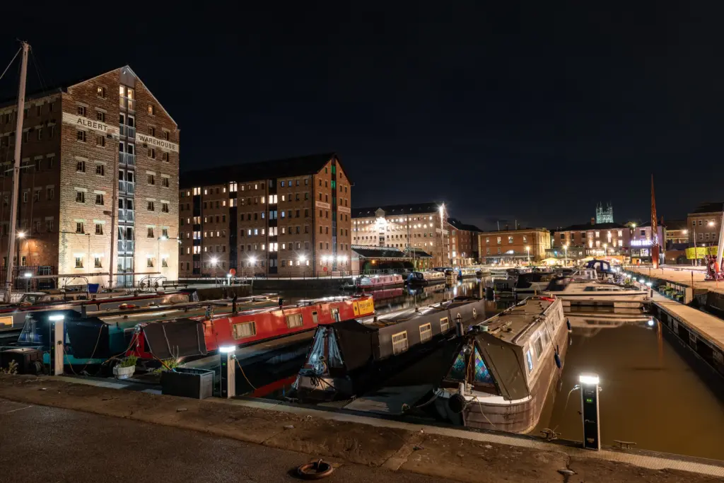 Albert Warehouse and docked canal boats at Gloucester Docks, with reflections in the water and a cathedral visible in the background at night.