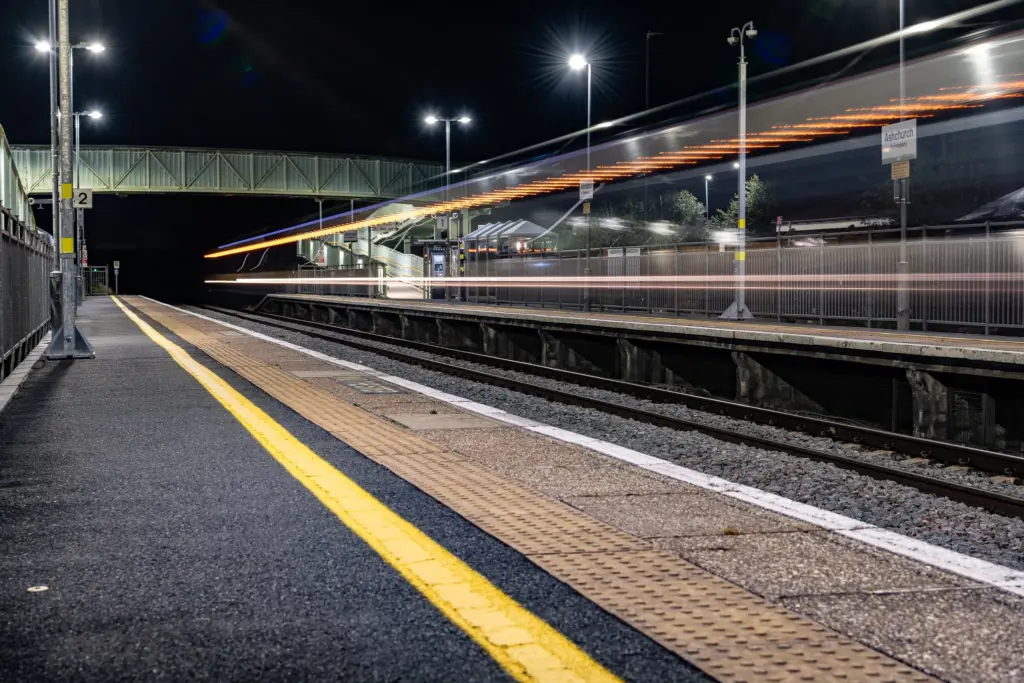 Nighttime view of Ashchurch for Tewkesbury railway station with a moving train, platform signage, and illuminated footbridge.