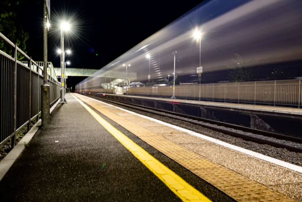Ashchurch for Tewkesbury train station at night with a motion-blurred train passing by and illuminated platform lights.