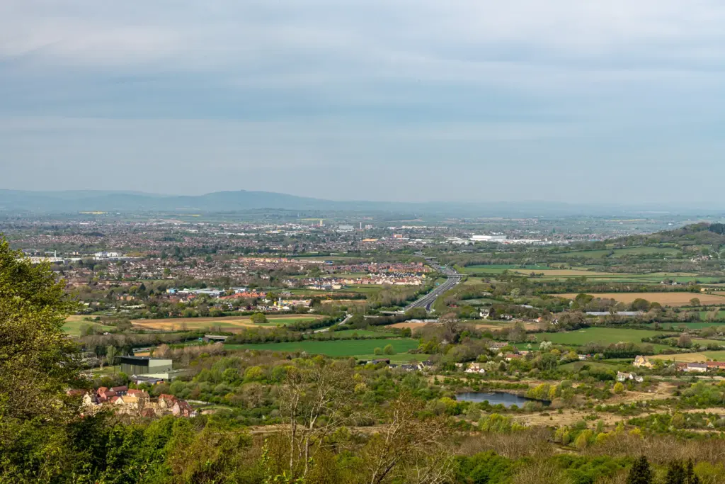 A panoramic view of Cheltenham with residential buildings, roads, and lush greenery, surrounded by hills under a cloudy sky.