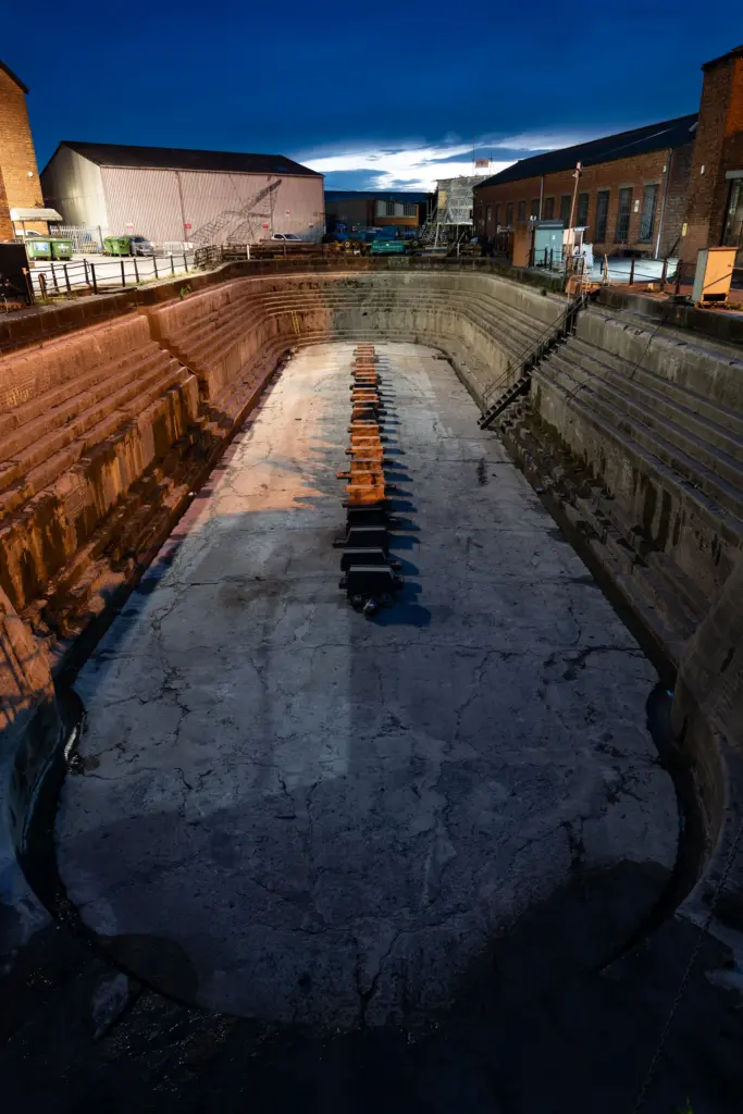 Empty dry dock at Gloucester Docks surrounded by brick and industrial buildings, captured at dusk with visible wooden blocks and stairs.
