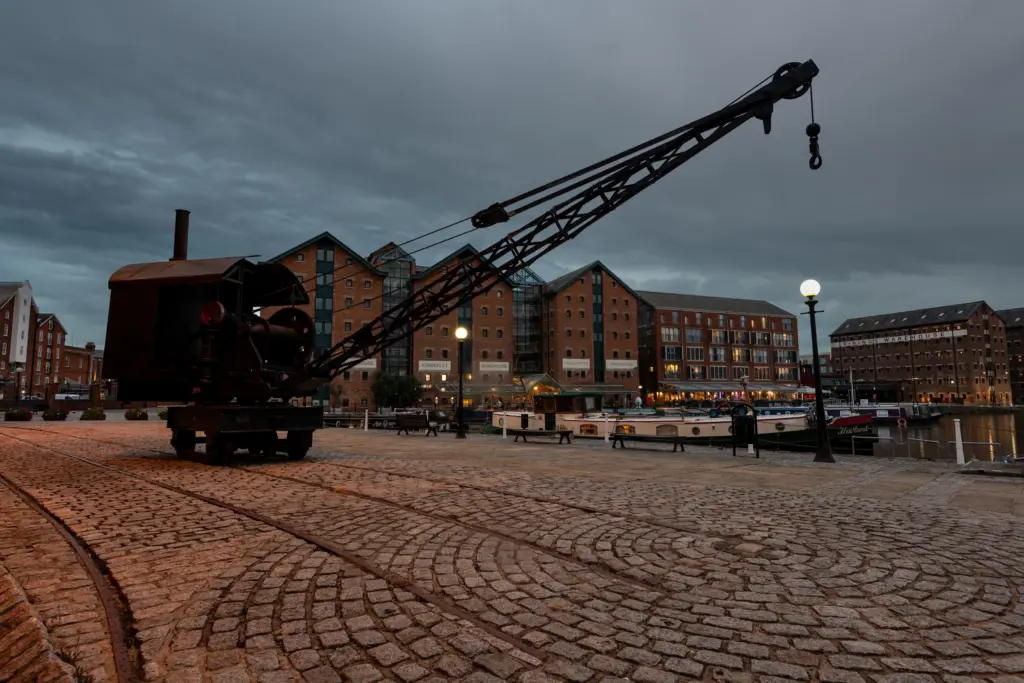 A historic Gloucester Docks waterfront scene with a large industrial crane, cobblestone pathway, docked canal boats, and brick warehouses under an overcast sky.