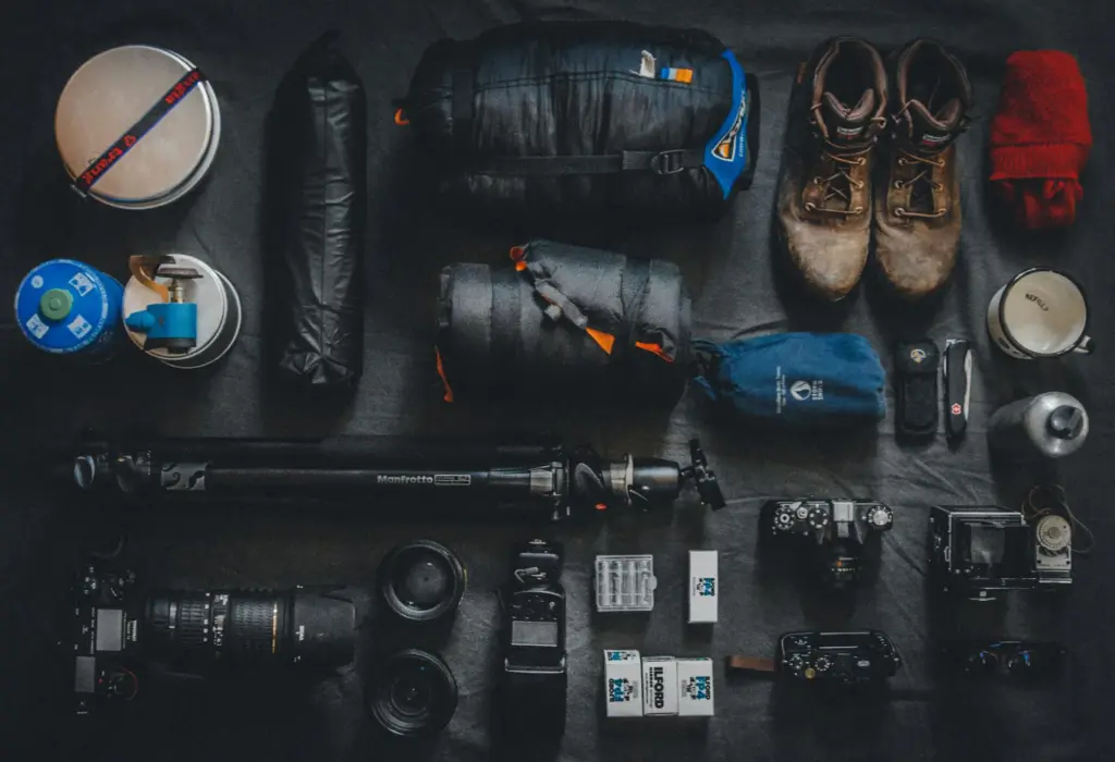 A flatlay of outdoor and photography gear, including a DSLR camera, lenses, tripod, hiking boots, camping stove, and film rolls, neatly arranged for an adventure.