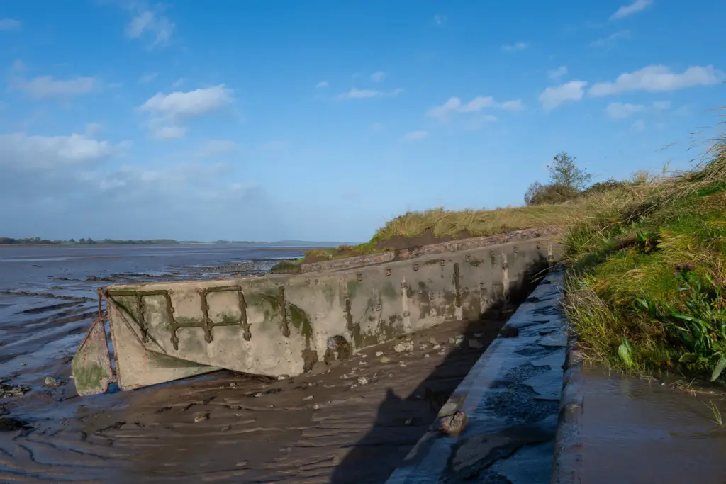Weathered concrete structure partially embedded in mud at Purton Ship Graveyard, surrounded by grassy banks and a calm waterbody under a blue sky
