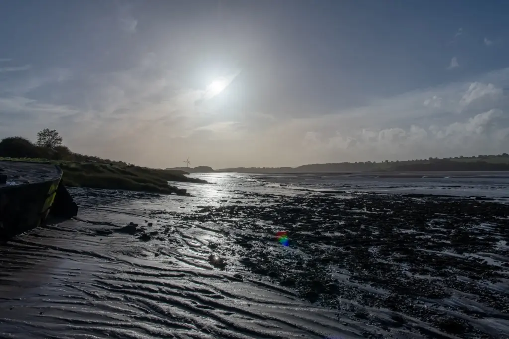 Coastal landscape at Purton Ship Graveyard during low tide, featuring mudflats, a wind turbine in the distance, and a rainbow reflection on wet sand