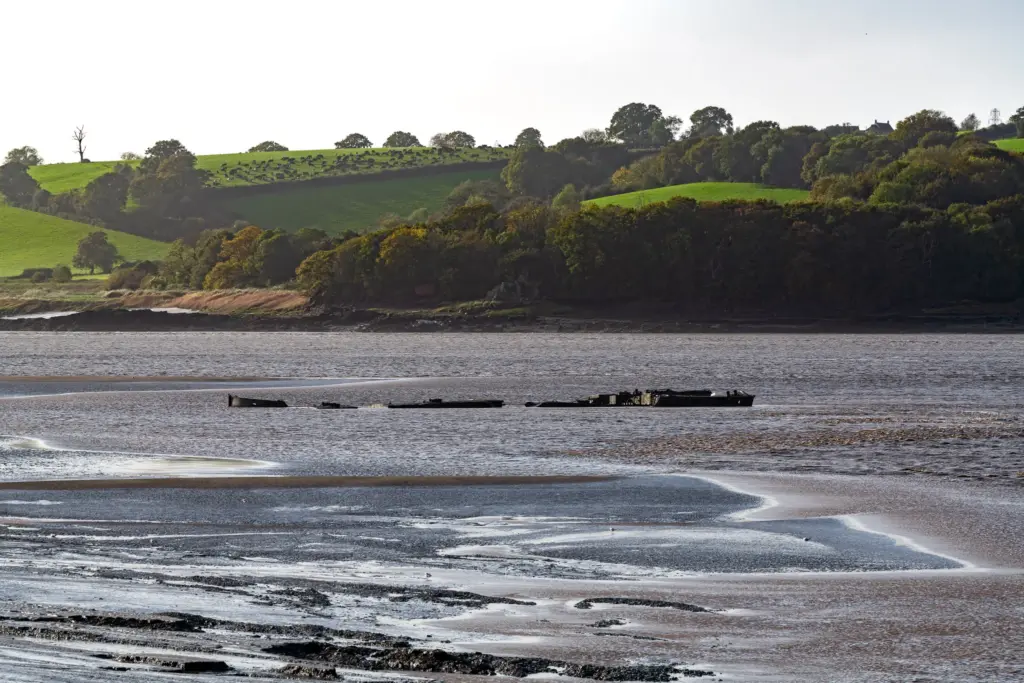 Partially submerged structure in a serene river at Purton Ship Graveyard, with rolling green hills and trees in the background