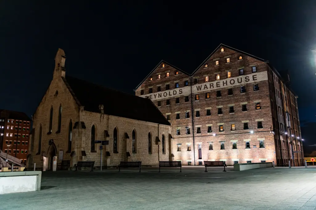 Reynolds Warehouse and a Gothic-style stone building at Gloucester Docks, illuminated at night with benches in the open plaza.