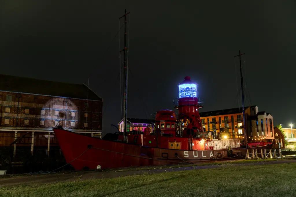The SULA lightship docked at Gloucester Docks at night, featuring a bright blue lighthouse and historic brick buildings in the background.