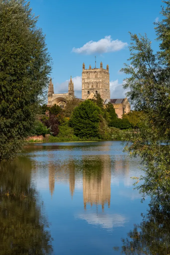 Tewkesbury Abbey surrounded by flood water, with its reflection visible in the calm surface, framed by lush greenery and a clear blue sky