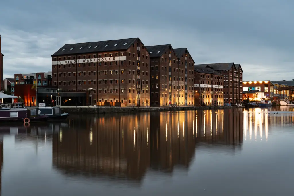 Twilight waterfront view of Vinings Warehouse at Gloucester Docks with illuminated brick buildings, calm water reflections, and moored boats.